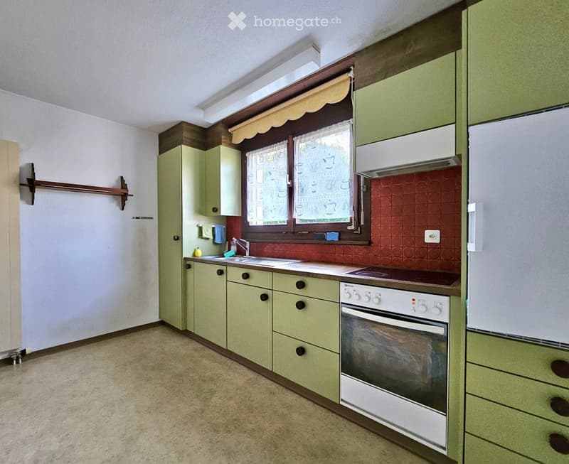 A small kitchen with green cabinets, a white oven, a sink, and a window with a decorative curtain. The backsplash is red with a circular pattern.