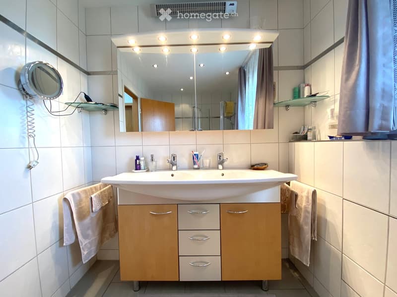 Modern bathroom with a double sink, large mirror with built-in lighting, wooden cabinet, and various toiletries.