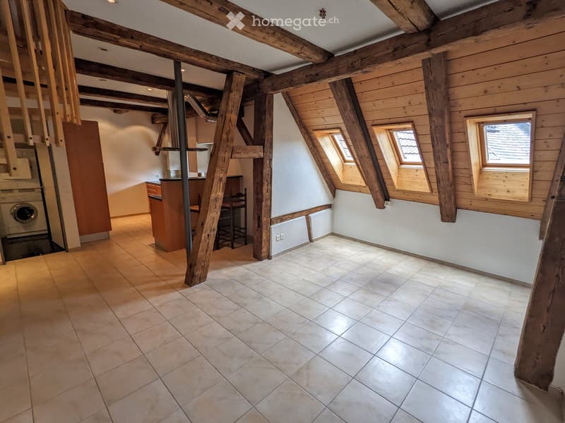 An unfurnished attic room with exposed wooden beams, tiled flooring, and skylight windows. The space includes a small kitchen area with cabinets and a washing machine, but no furniture is present.