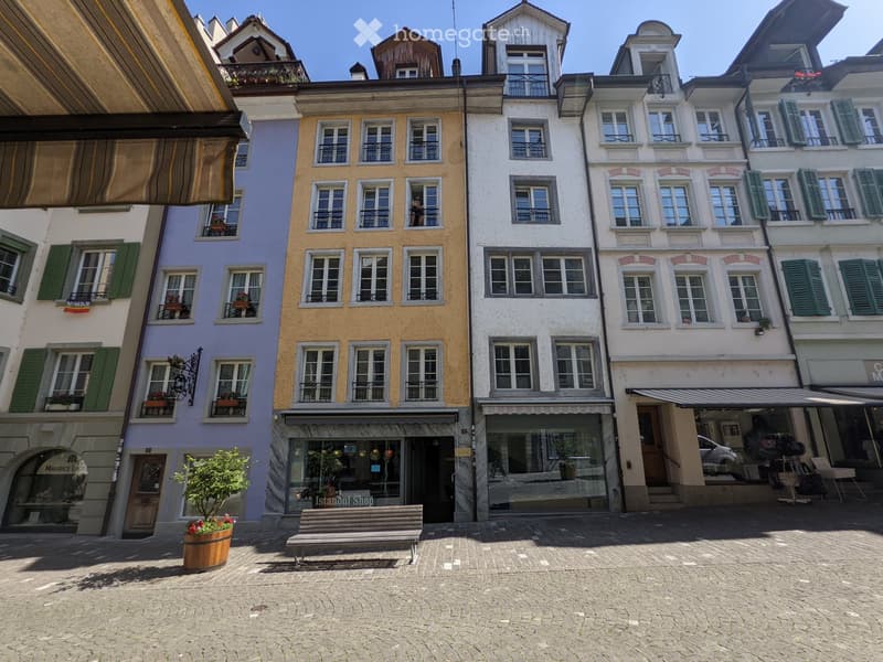 A row of colorful multi-story buildings with green shutters, balconies, and storefronts, including a bench and a small tree in a barrel on the cobblestone street.