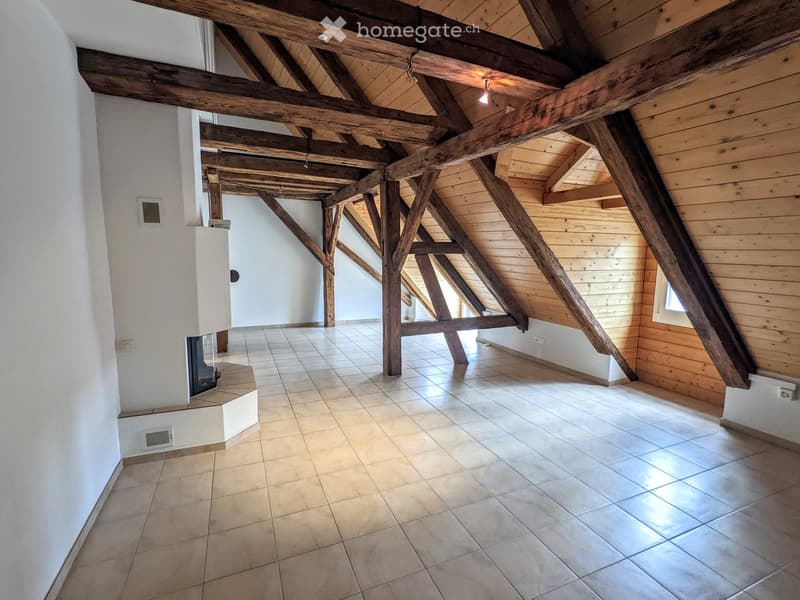 An empty attic space with exposed wooden beams, tiled floor, and a small fireplace area. The room has white walls, wooden paneling on one side, and a skylight window.