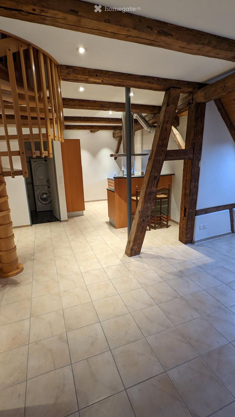 Loft-style room with exposed wooden beams, tiled floor, staircase leading to an upper level, and a small kitchen area with a washing machine and seating.