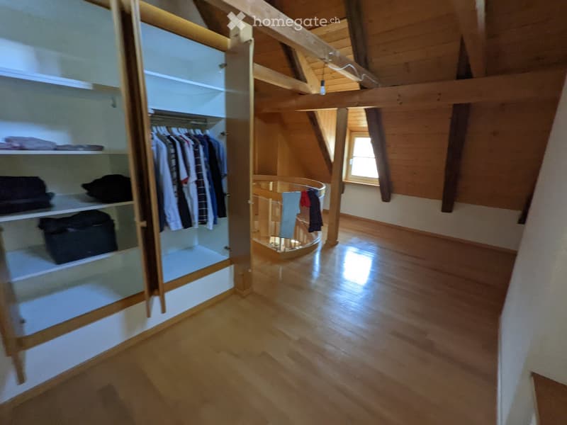 Spacious attic room with wooden floors, exposed wooden beams, and large windows. Features a built-in wardrobe with shelves and hanging space, and a small round table with a few folded clothes on it.