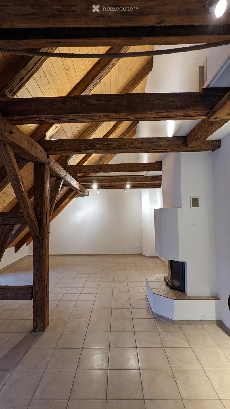 An empty room with exposed wooden beams and a tiled floor, featuring a small built-in fireplace and recessed lighting.