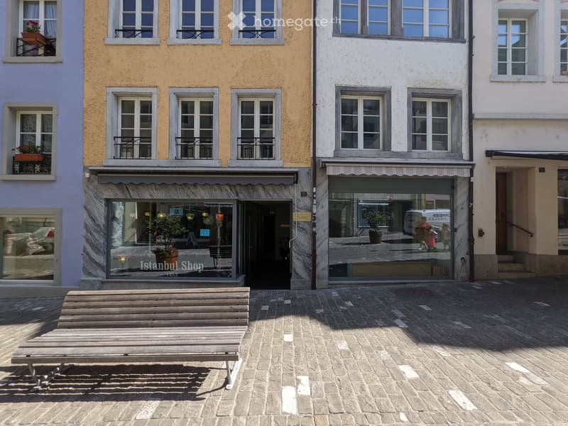 A paved outdoor area with a wooden bench, surrounded by colorful buildings with balconies and shopfronts.