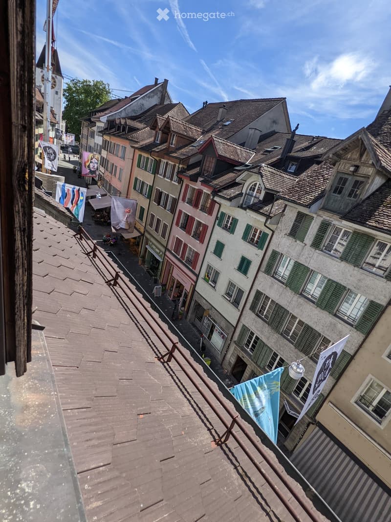 View from a rooftop terrace overlooking a historic European town with colorful buildings, tiled roofs, and narrow streets.