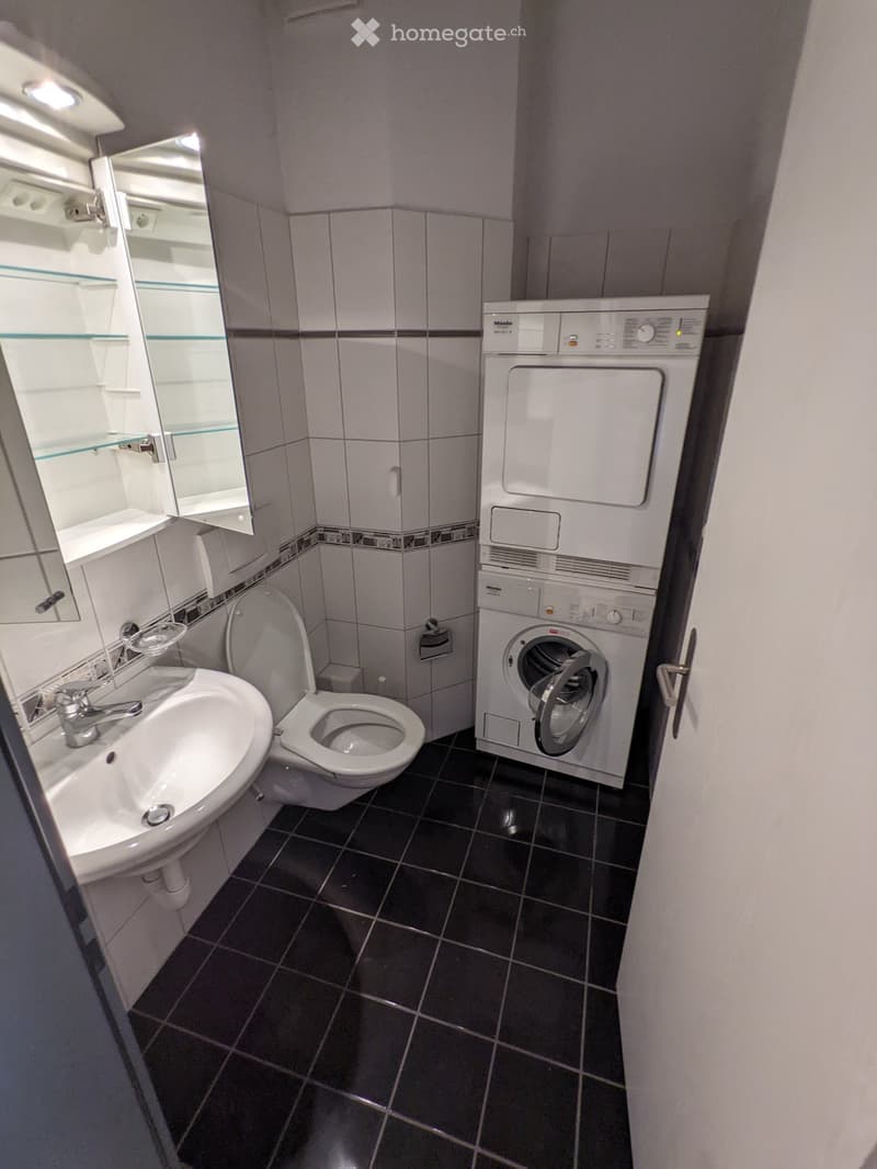 Modern bathroom with dark tiled floor, white sink, toilet, mirror, and a stacked washer/dryer unit.