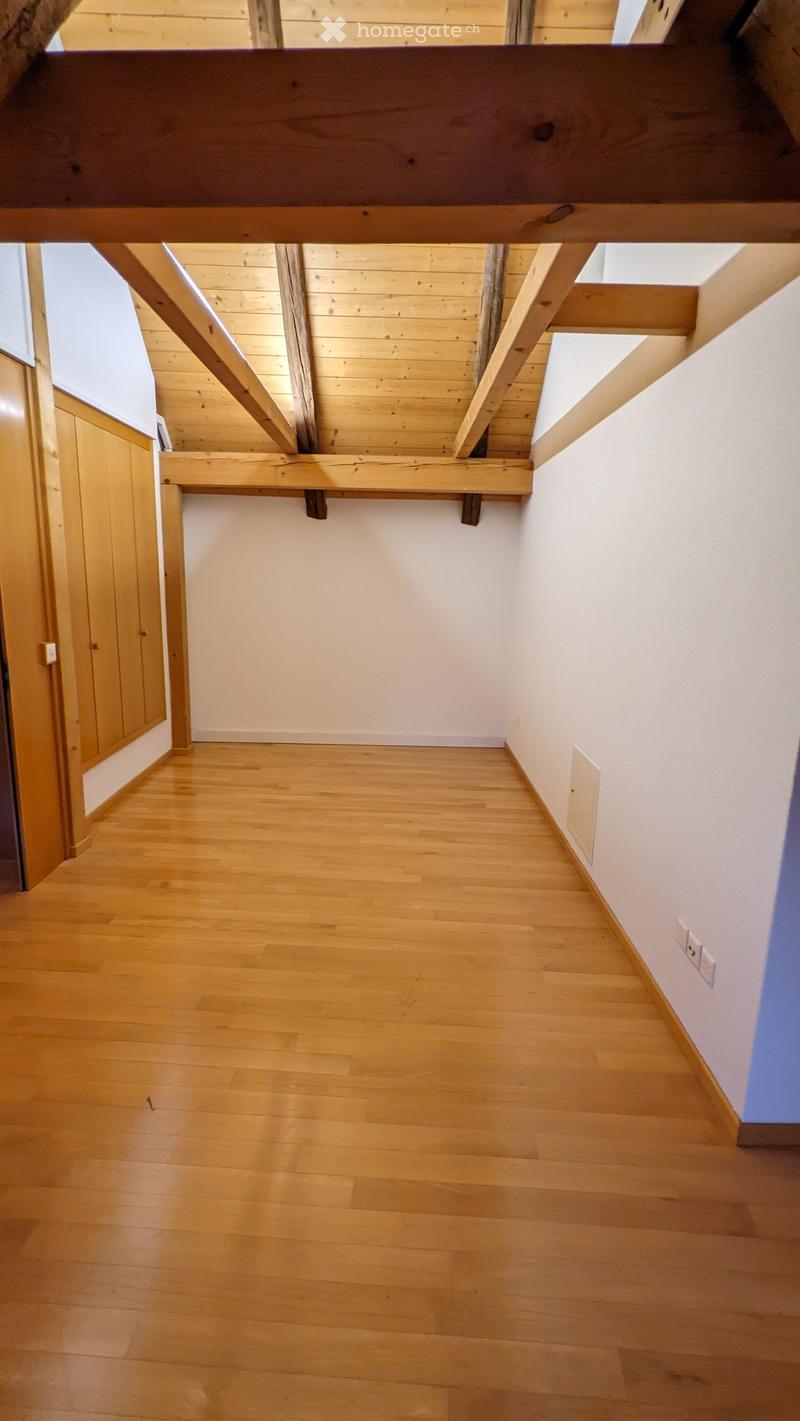 Empty hallway with wooden flooring, white walls, and exposed wooden beams on the ceiling. Features a wooden cabinet on the left and a small window at the far end.