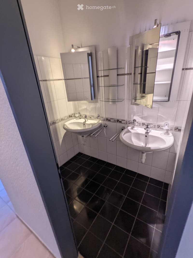 Modern bathroom with two sinks, mirrors, and glass shelves above them. Dark tiled floor and white tiled walls.