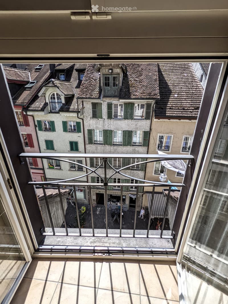 Balcony overlooking a historic urban area with traditional buildings, green shutters, and a small courtyard below.