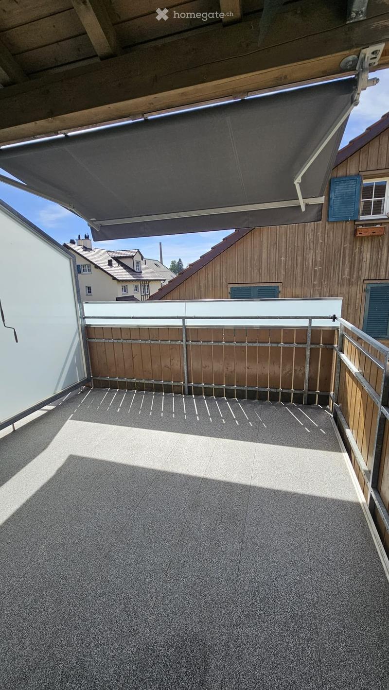 Roof terrace with a modern metal railing, wooden privacy screen, and a retractable awning. The floor is covered with a textured gray material, and the surrounding area includes wooden walls and neighboring buildings.