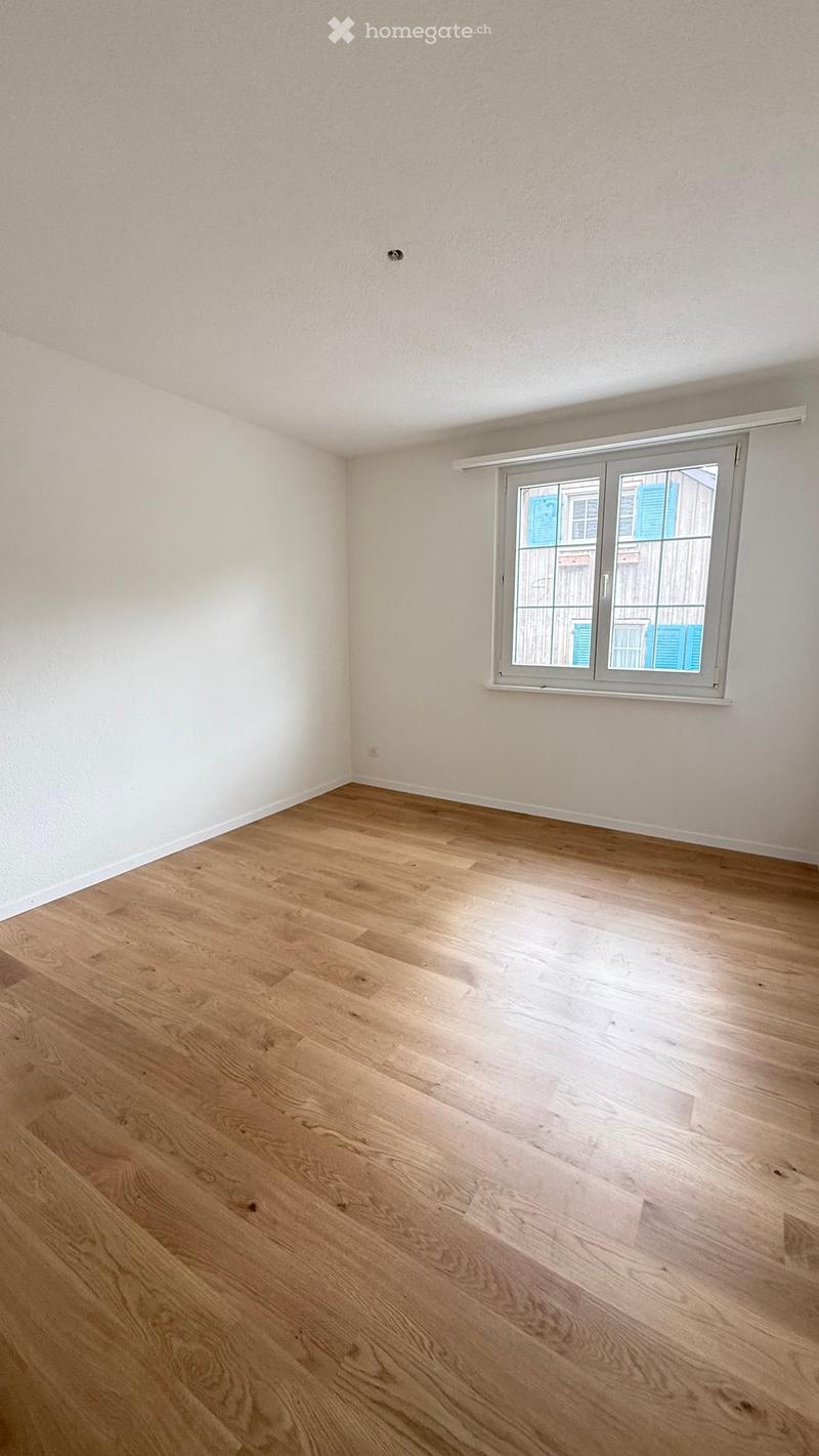 Empty room with light wooden flooring and white walls, featuring a large window with a view of neighboring buildings.
