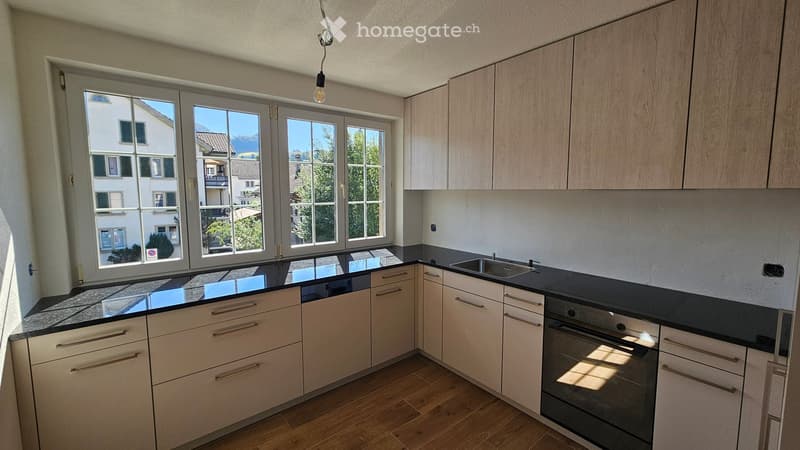 Modern kitchen with light wood cabinets, black countertops, and large windows overlooking a residential area.