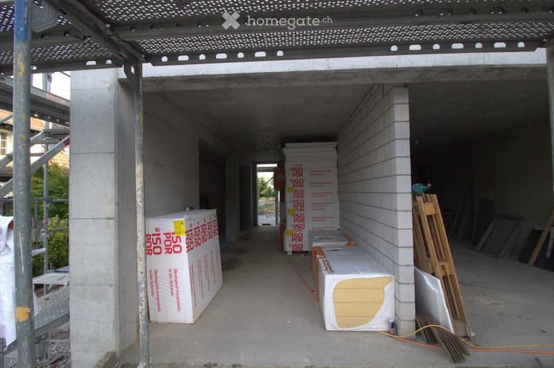 An under-construction corridor with exposed concrete floors and walls, metal roofing, and construction materials stacked along the sides.
