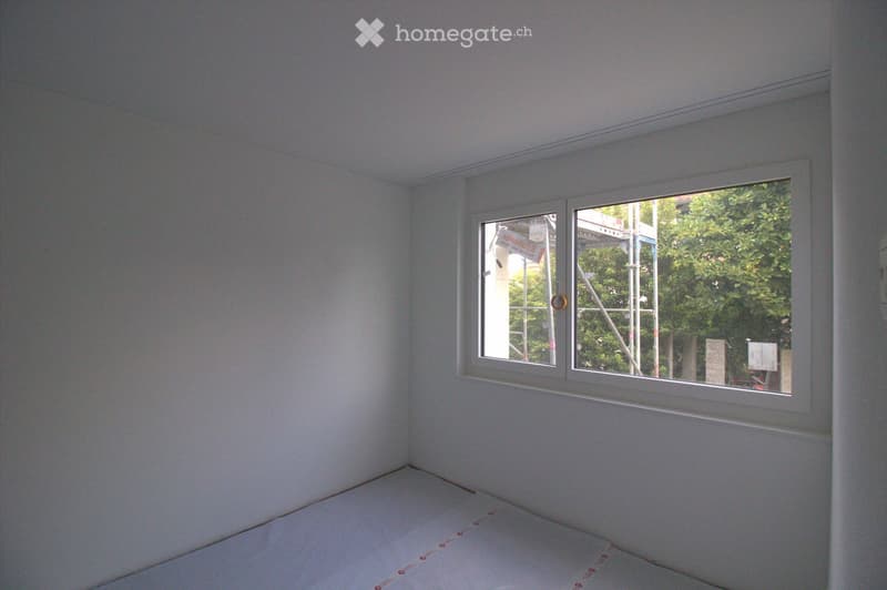Empty room with white walls and a large window showing a view of construction scaffolding and greenery outside.