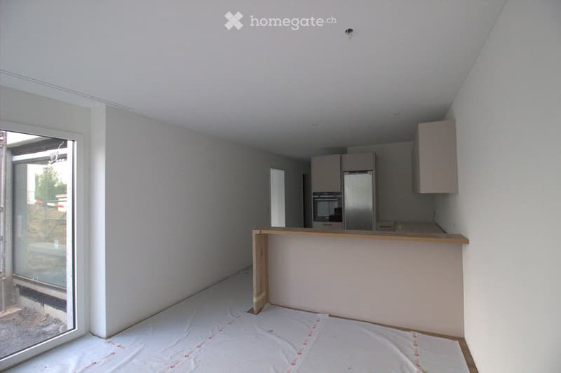 Empty room with white walls, wooden floor, and a large window with a view of the outside. The room has a kitchen area with cabinets and a refrigerator, but no furniture or appliances are present.