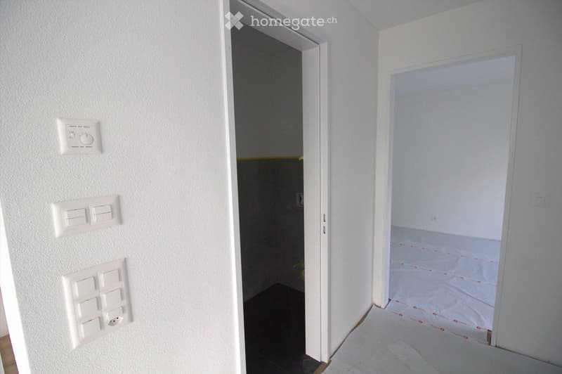 Empty room with white walls, light switches, and a partially visible adjacent room with a dark floor.