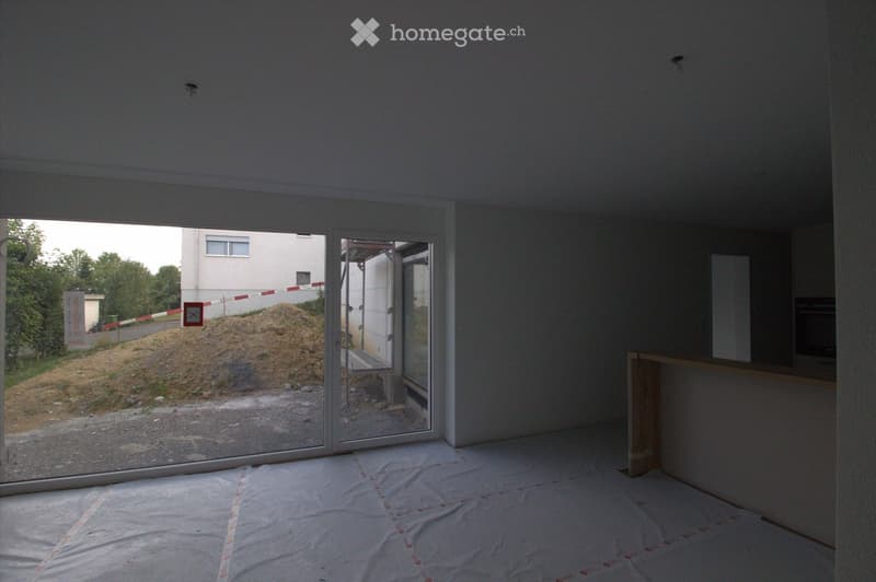 Empty room with bare walls and a wooden floor, covered with protective plastic sheeting. Large glass sliding doors lead to an outdoor area with a pile of dirt and construction materials.