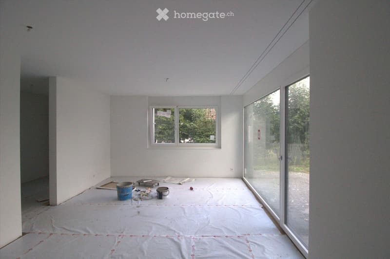 Empty room with white walls, wooden floor, and large windows, covered with protective plastic sheeting and some construction materials.