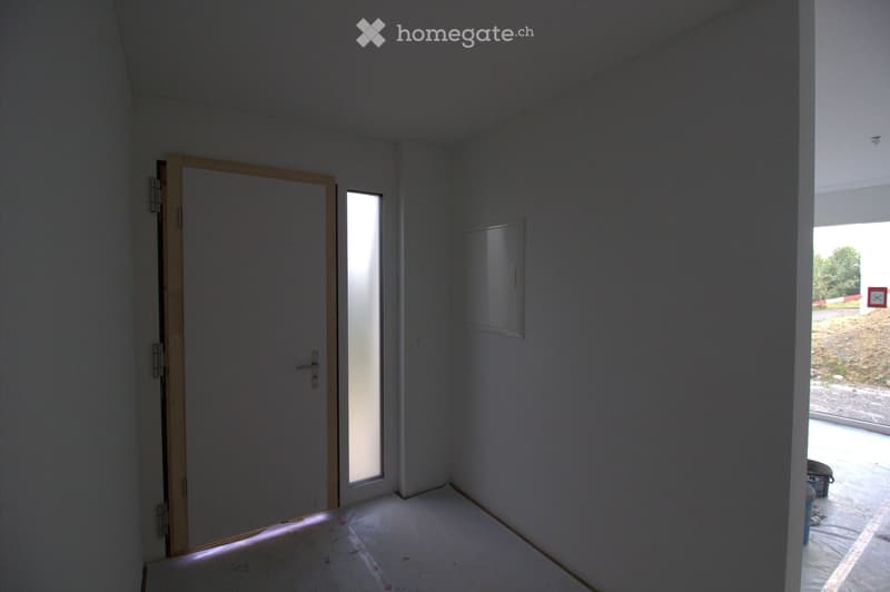 Empty room with white walls, a wooden door, a small window, and a partially visible exterior area through an open doorway.