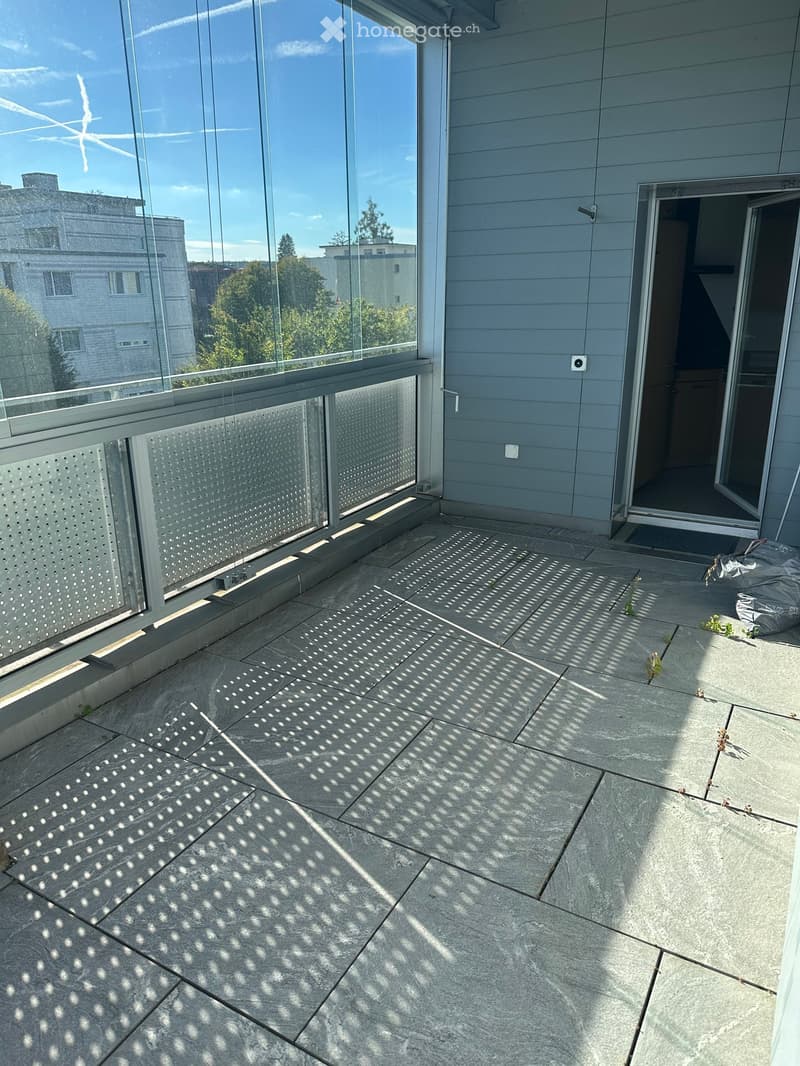 Modern balcony with large glass windows, grey tiled floor, and a view of surrounding buildings and greenery.