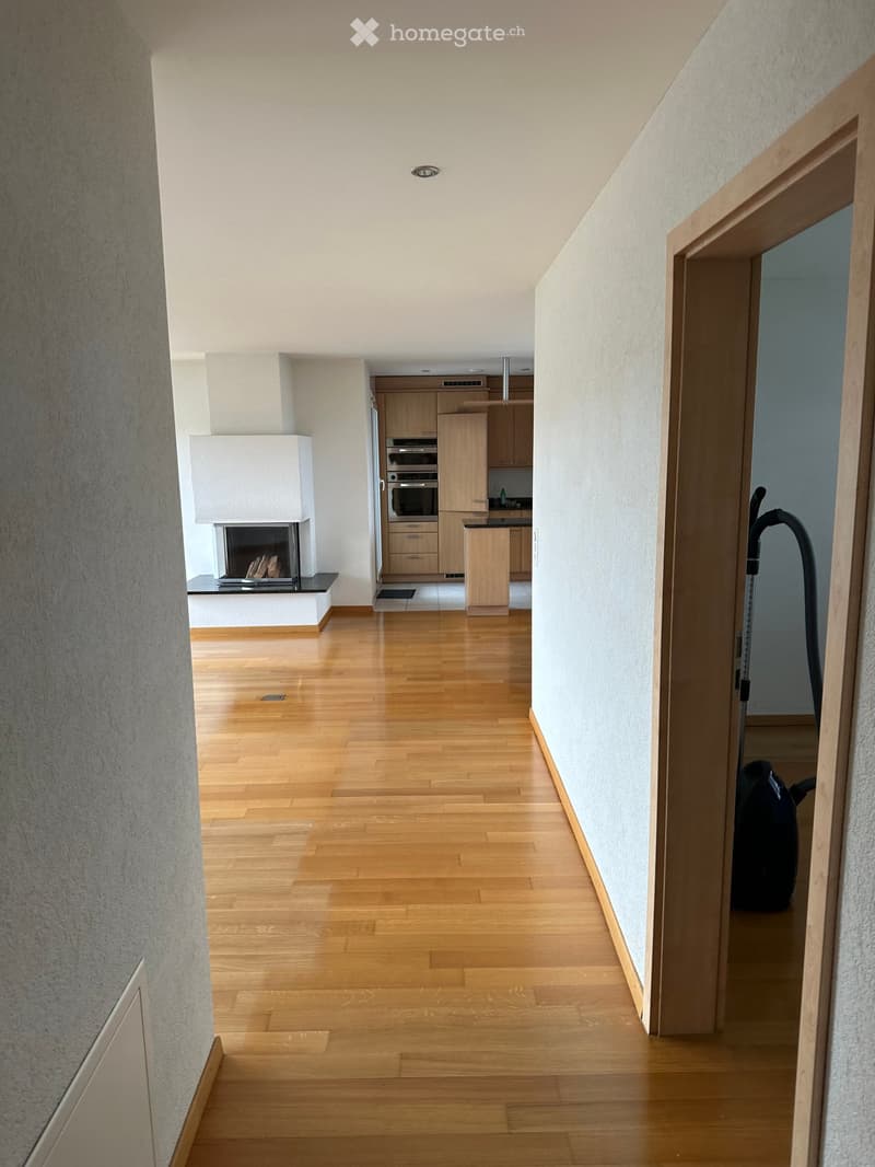 Spacious hallway with wooden flooring, white walls, and a fireplace on the left side. A kitchen area is visible in the background with wooden cabinets and stainless steel appliances.