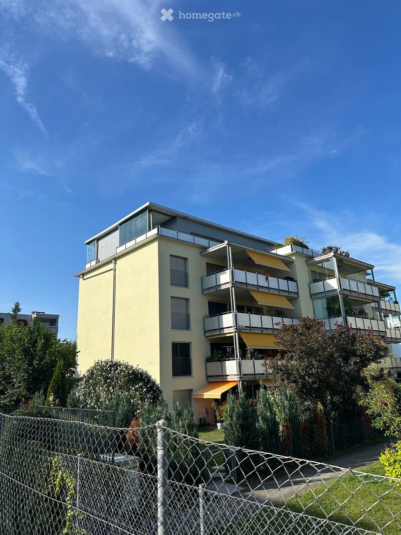 Modern multi-story residential building with balconies, large windows, and surrounding greenery.