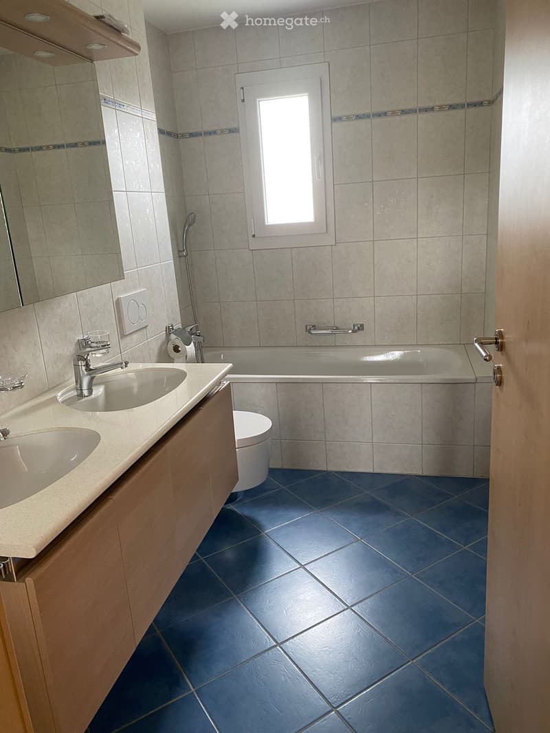 Bathroom with blue tiled floor, beige tiled walls, double sink vanity, bathtub, toilet, and a window above the bathtub.