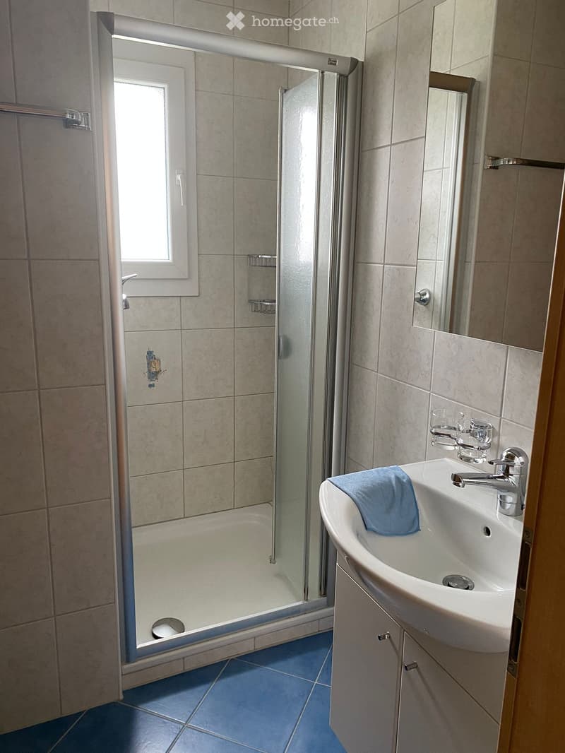 Bathroom with a white sink, blue towel, mirror, bathtub, and glass shower enclosure. Blue tiles on the floor and beige tiles on the walls.