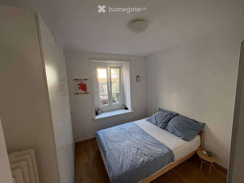Cozy bedroom with a single bed, blue bedding, a small nightstand, and a wooden stool. The room features light-colored walls, wooden flooring, and a window with a view of the outside.