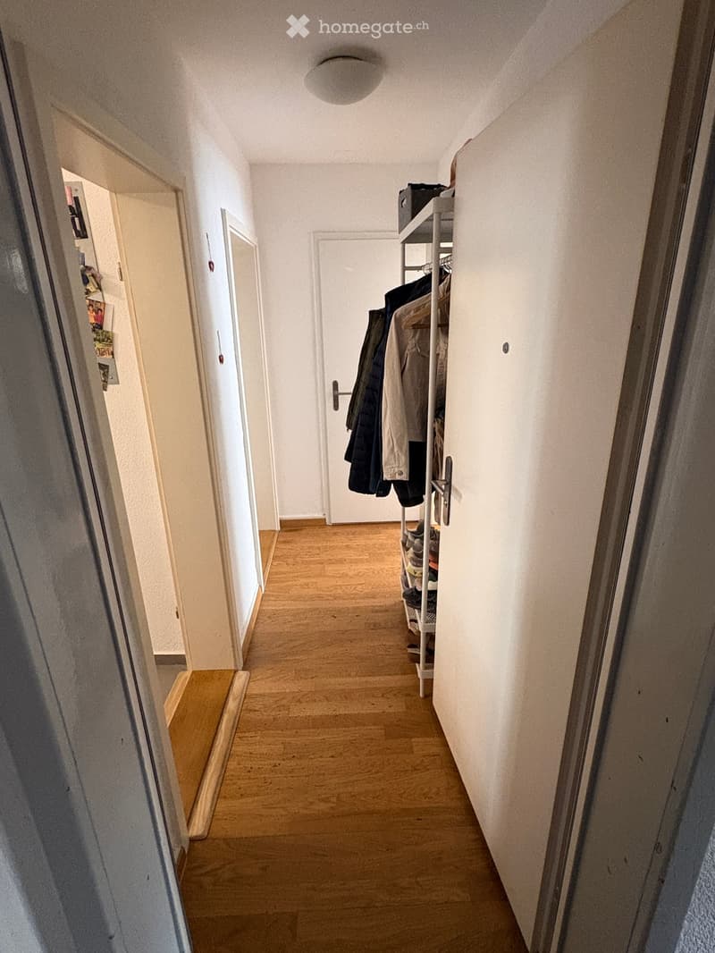A narrow hallway with wooden flooring, white walls, and a coat rack holding jackets and shoes. The hallway leads to an open door at the end.