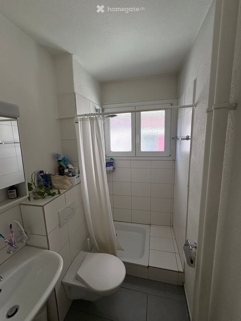 A small bathroom with a toilet, bathtub, sink, and mirror. The space includes a shower curtain, a toothbrush holder, and a cabinet with toiletries.