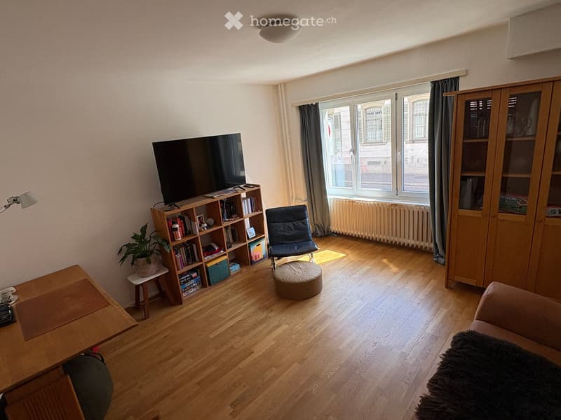 Living room with wooden flooring, large window with curtains, sofa, coffee table, bookshelf, chair, and television.