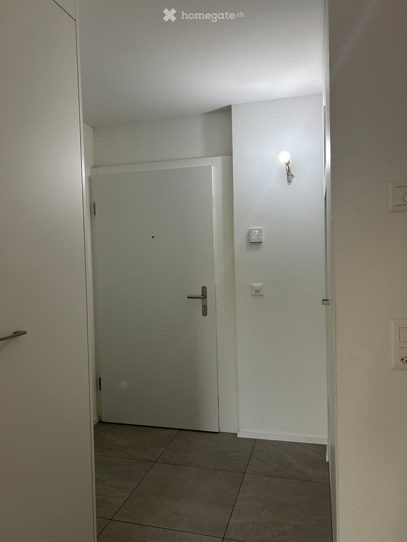 Empty hallway with tiled floor, white walls, and a closed door. Light fixture mounted on the wall and electrical outlets visible.