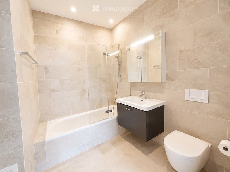 Modern bathroom with beige tiles, a bathtub, a sink with a dark cabinet, a toilet, and a large mirror with built-in lighting.