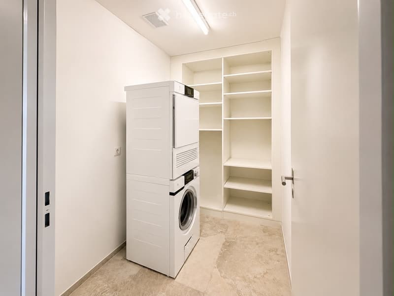 A clean, modern laundry room with white walls, tiled floor, and built-in shelves. Features a stacked washer and dryer unit.