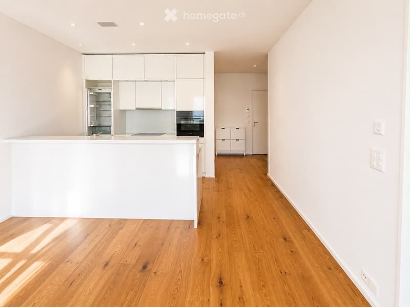 Modern kitchen with white cabinetry, a central island, and wooden flooring. Features an oven and refrigerator, with recessed lighting and a hallway leading to another room.
