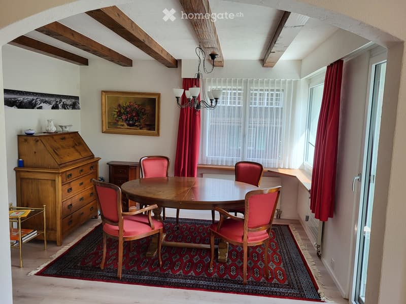 Dining room with wooden dining table, six chairs with red cushions, wooden cabinet, chandelier, painting, and red curtains.