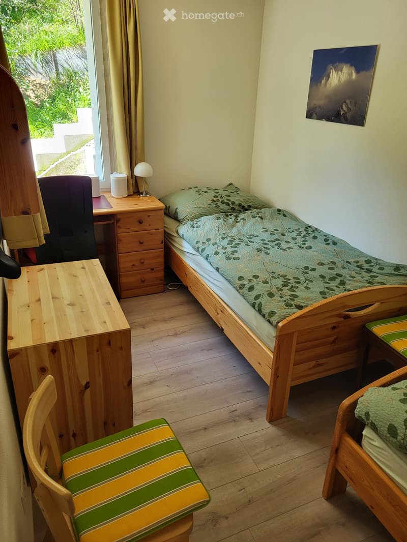 Cozy bedroom with wooden bed, small desk, chair, and a window with curtains. Features a painting on the wall and a small table with a lamp.