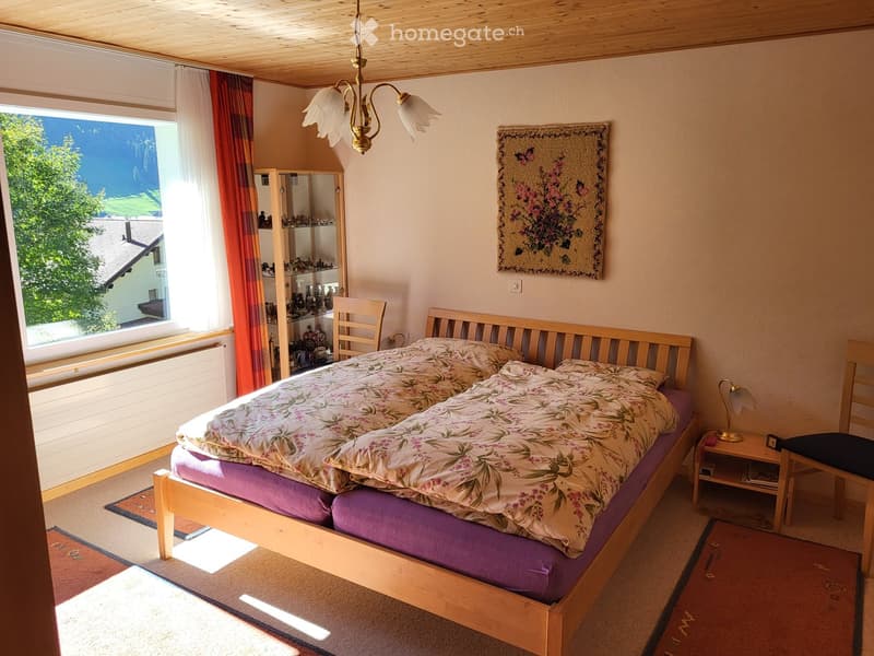 Cozy bedroom with wooden bed, floral duvet, nightstand, chair, mirror cabinet, and wall tapestry. Large window with view of greenery.