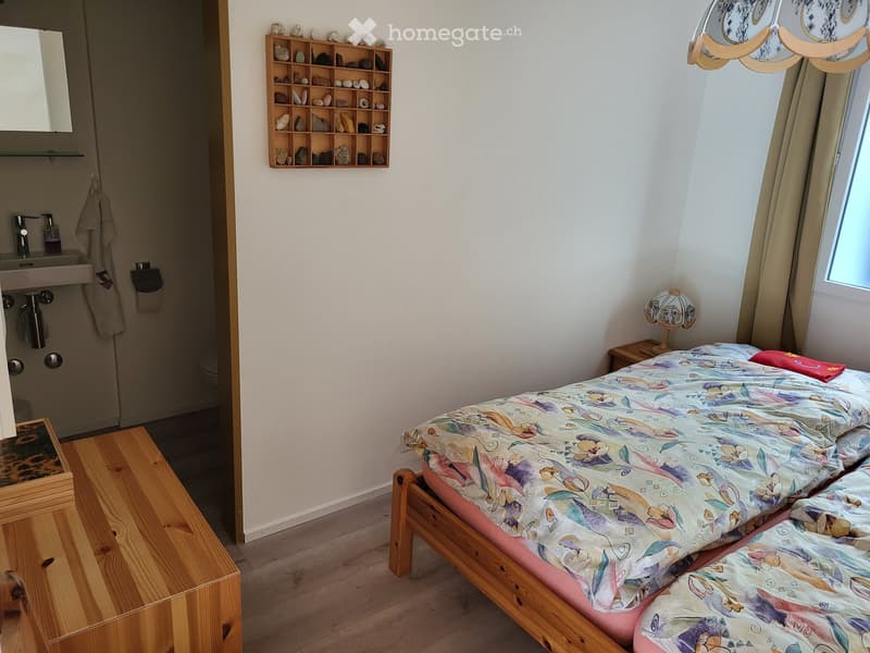 Cozy bedroom with a wooden bed, colorful bedspread, nightstand with lamp, small sink area with mirror, and a wooden shelf with decorative stones.