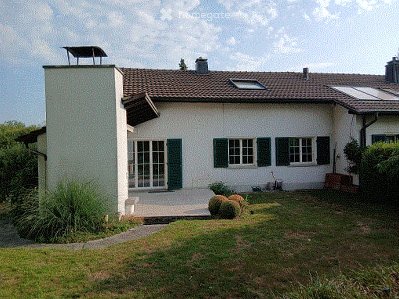 Single-story house with white exterior, brown tiled roof, multiple windows with green shutters, small garden area with shrubs and a lawn.