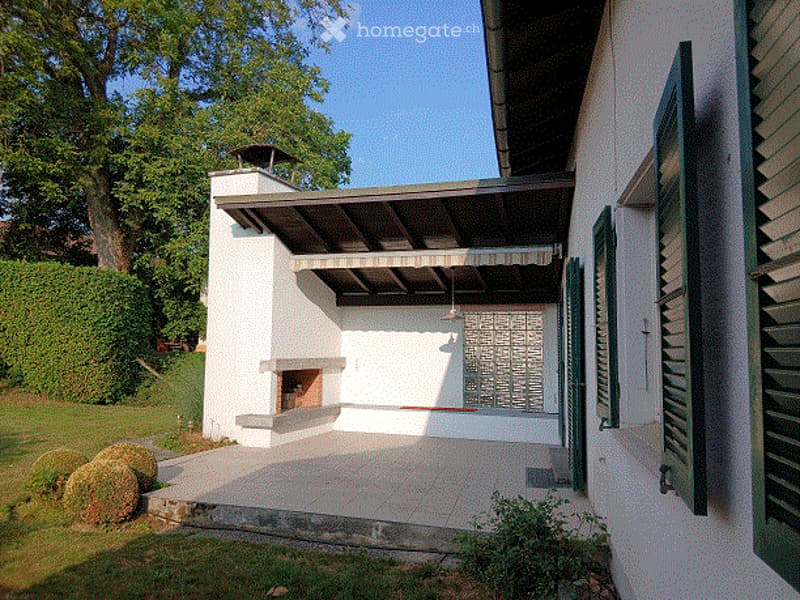 Modern outdoor patio area attached to a white house, featuring a built-in fireplace, large green shutters, and a covered seating area with a slatted roof.