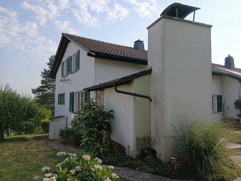 A two-story white house with green shutters, a tiled roof, and a chimney. The house is surrounded by a well-maintained garden with flowering plants and trees.