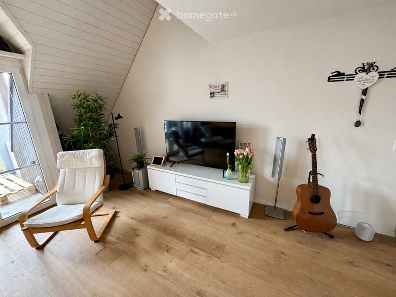 Modern living room with wooden flooring, white walls, and slanted ceiling. Features a white armchair, a guitar, a television on a white cabinet, a vase with flowers, and speakers.