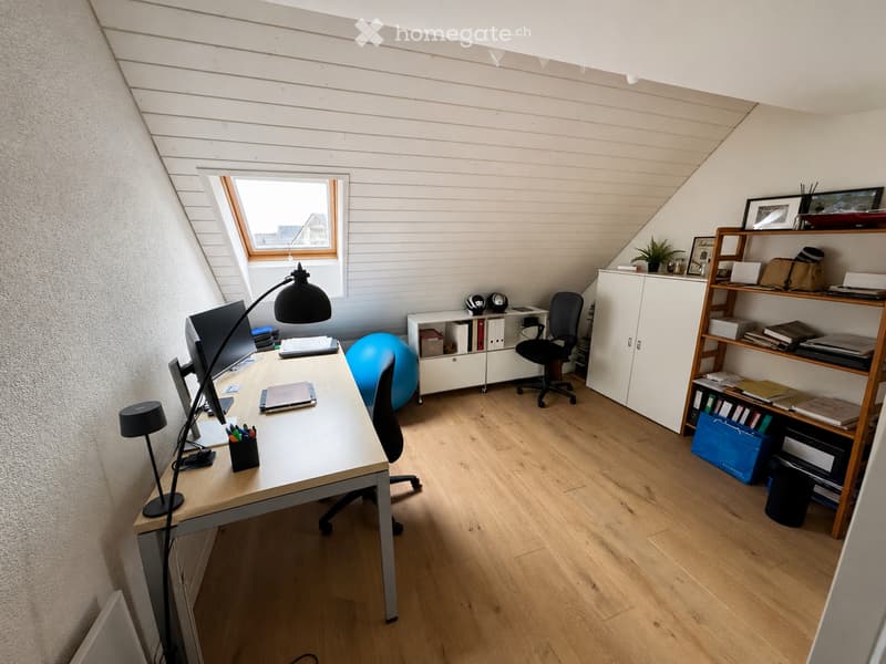 A modern, well-lit office space with wooden flooring, a large window, a desk with a computer, a chair, a blue exercise ball, and shelves with books and office supplies.