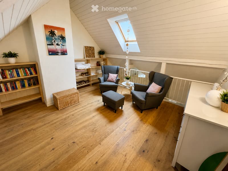 Cozy attic room with wooden flooring, two armchairs, a small table, bookshelf, and decorative items.