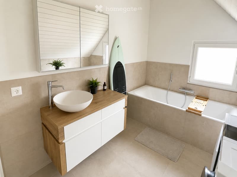 Modern bathroom with a wooden vanity, white sink, bathtub, large mirror, and a surfboard leaning against the wall.