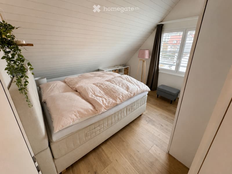Cozy attic bedroom with wooden flooring, a large bed with pink bedding, a floor lamp, a small nightstand, and a decorative plant on a shelf.