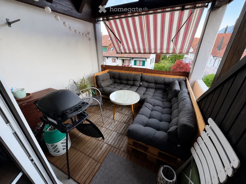 Balcony with wooden decking, grey sectional sofa, round table, white chairs, barbecue, fan, and potted plant.